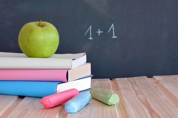 Three books with different colored covers lie on a wooden surface in front of a blackboard with an apple - concept for education and school with place for text or other elements