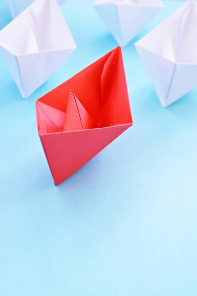 A red self-folded paper boat lies on a blue surface, followed by many white boats - concept symbolizing leadership