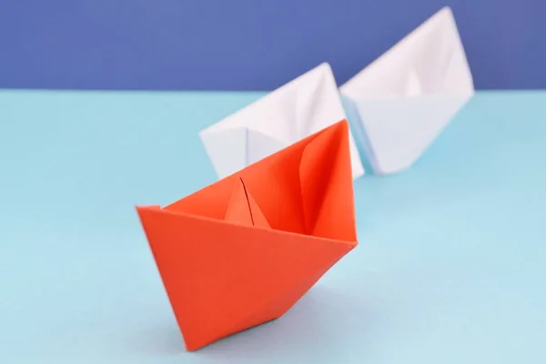 A red self-folded paper boat lies on a blue surface, followed by many white boats - concept symbolizing leadership