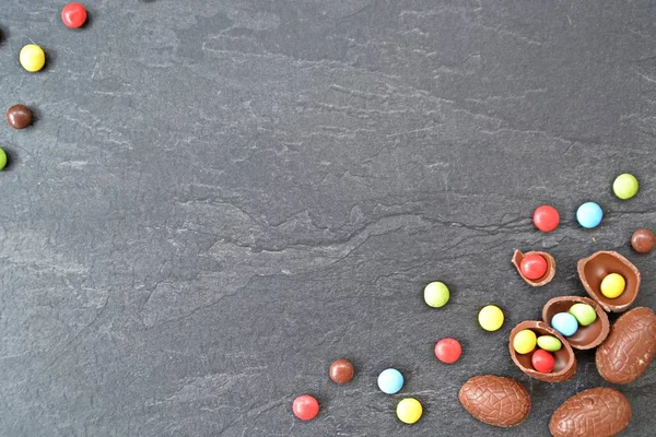 A few chocolate eggs on a dark marble surface, some of which are broken, in the eggs and next to it are colorful chocolate - concept with Easter sweets and space for text or other elements