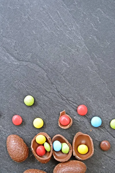 A few chocolate eggs on a dark marble surface, some of which are broken, in the eggs and next to it are colorful chocolate - concept with Easter sweets and space for text or other elements