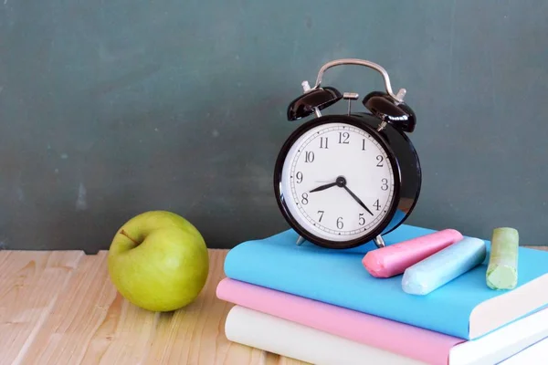 An alarm clock stands in front of a blackboard with colorful books and a green apple - concept with space for text or other elements on the subject of school and time management in elementary school