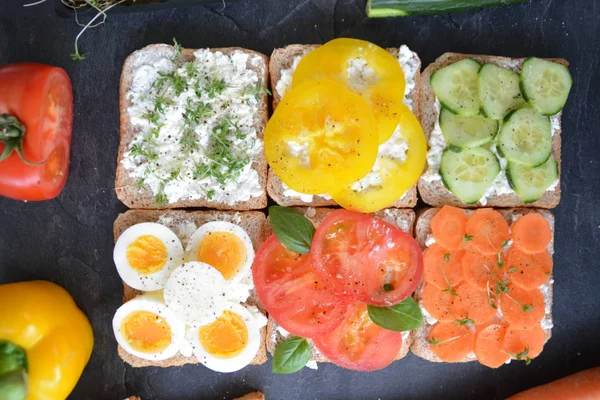 Many whole-grain toasts with different toppings lie on a dark surface - concept for healthy nutrition with vegetables on a wholemeal toast and coarse-grained cream cheese as background