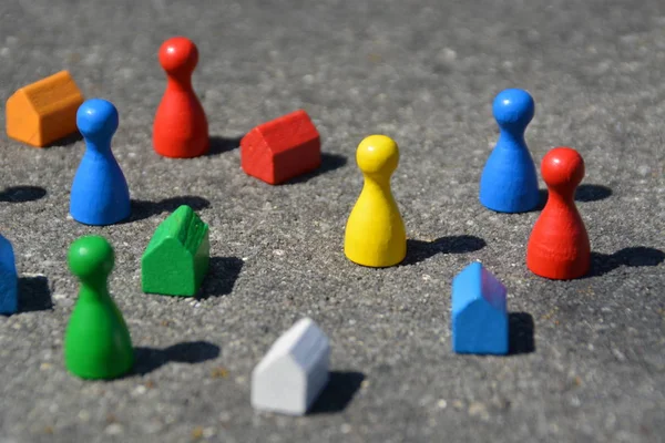 Differently colored game figures stand together with cubes and small houses in different colors on a stone basis and cast by the high sun a shadow