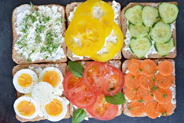Many whole-grain toasts with different toppings lie on a dark surface - concept for healthy nutrition with vegetables on a wholemeal toast and coarse-grained cream cheese as background