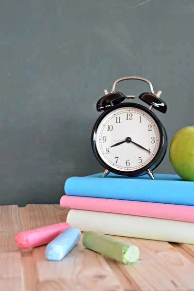 An alarm clock stands in front of a blackboard with colorful books and a green apple - concept with space for text or other elements on the subject of school and time management in elementary school