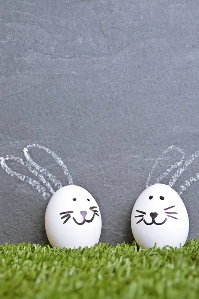 A white egg with a rabbit face painted on it lies on a lawn in front of a blackboard on which bunny ears were painted - Easter concept with space for text or other elements