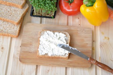 Kaba-taneli krem peyniri ve Cress ile bir kepekli tost bir bıçak yanında bir tahta tahta ve biber ve domates gibi sebze, yanı sıra Cress, salatalık ve havuç bir masada yatıyor