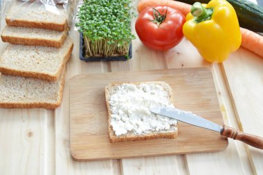 Kaba-taneli krem peyniri ve Cress ile bir kepekli tost bir bıçak yanında bir tahta tahta ve biber ve domates gibi sebze, yanı sıra Cress, salatalık ve havuç bir masada yatıyor