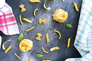 Pasta noodles of the sort tagliatelle lie on a dark marble surface which is lightly sprinkled with flour, next to it are basil leaves - concept as background for fresh prepared noodles clipart