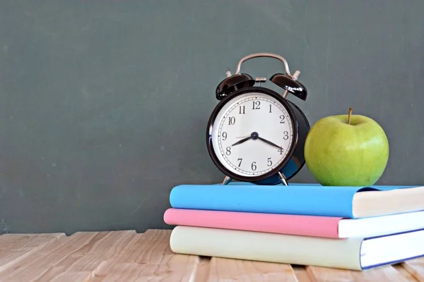 An alarm clock stands in front of a blackboard with colorful books and a green apple - concept with space for text or other elements on the subject of school and time management in elementary school