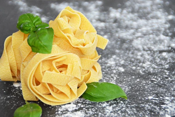 Pasta noodles of the sort tagliatelle lie on a dark marble surface which is lightly sprinkled with flour, next to it are basil leaves - concept as background for fresh prepared noodles