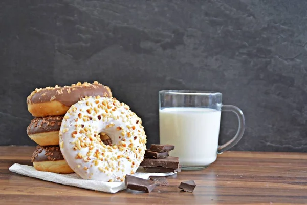 glazed donut with brittle splinters and other chocolate donuts lie on a dark wooden surface against a dark background and a glass of milk - concept for sweet donuts