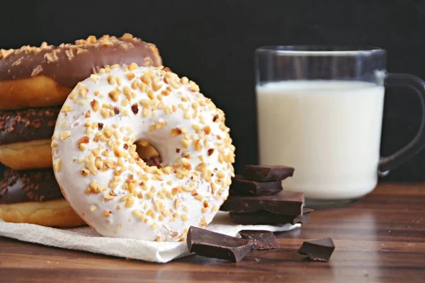 glazed donut with brittle splinters and other chocolate donuts lie on a dark wooden surface against a dark background and a glass of milk - concept for sweet donuts