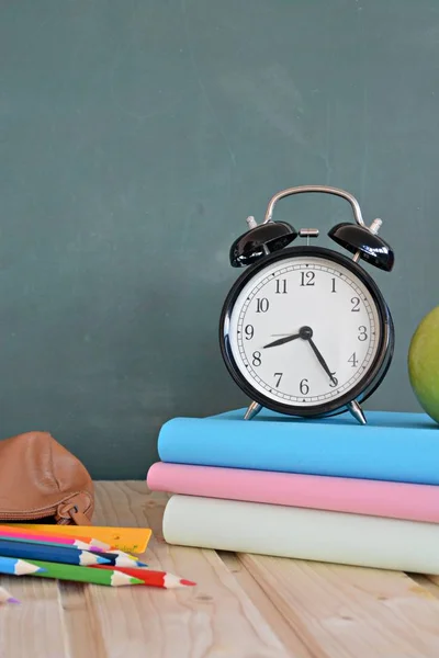 An alarm clock stands in front of a blackboard with colorful books and a green apple - concept with space for text or other elements on the subject of school and time management in elementary school