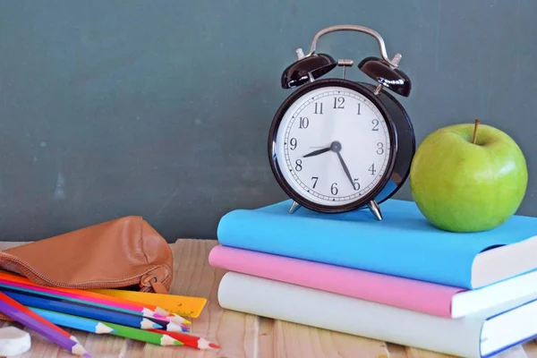 An alarm clock stands in front of a blackboard with colorful books and a green apple - concept with space for text or other elements on the subject of school and time management in elementary school