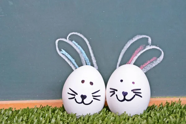 A white egg with a rabbit face painted on it lies on a lawn in front of a blackboard on which bunny ears were painted - Easter concept with space for text or other elements