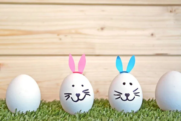 eggs painted with the face of a hare and glued to rabbit ears lie on a grass field in front of a wooden background with space for text or other elements