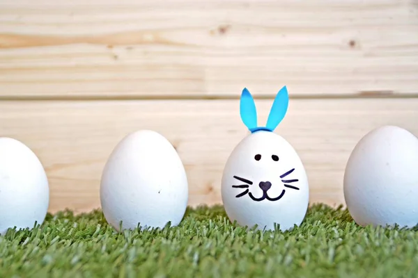 eggs painted with the face of a hare and glued to rabbit ears lie on a grass field in front of a wooden background with space for text or other elements