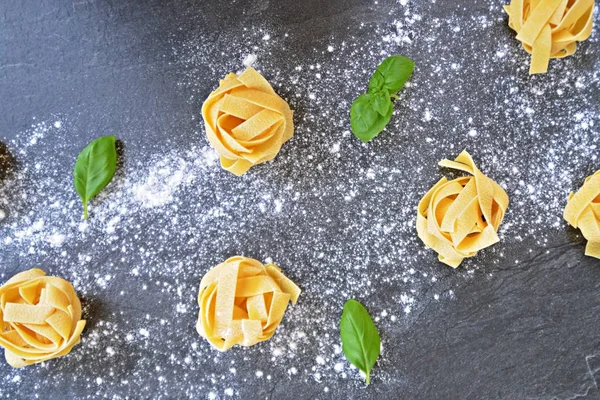 Pasta noodles of the sort tagliatelle lie on a dark marble surface which is lightly sprinkled with flour, next to it are basil leaves - concept as background for fresh prepared noodles