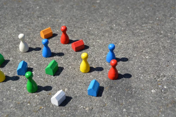 Differently colored game figures stand together with cubes and small houses in different colors on a stone basis and cast by the high sun a shadow