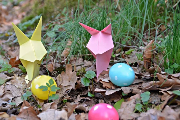 Bunnies folded from colorful paper in origami style stand with a few colorful plastic eggs with white spots on a forest floor in spring