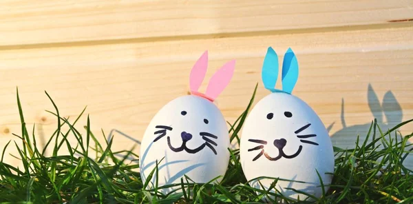 eggs painted with the face of a hare and glued to rabbit ears lie on a grass field in front of a wooden background with space for text or other elements