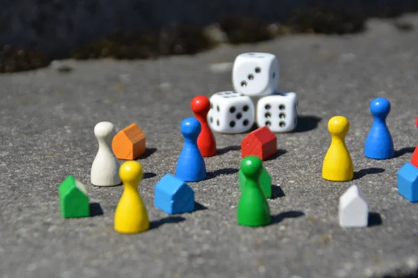 Differently colored game figures stand together with cubes and small houses in different colors on a stone basis and cast by the high sun a shadow