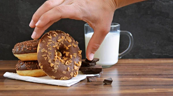 glazed donut with brittle splinters and other chocolate donuts lie on a dark wooden surface against a dark background and a glass of milk - concept for sweet donuts