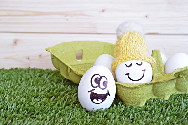 Two eggs with funny faces painted on it lie on a grass field. One of the eggs is in an egg box with other white eggs, the other is next to it