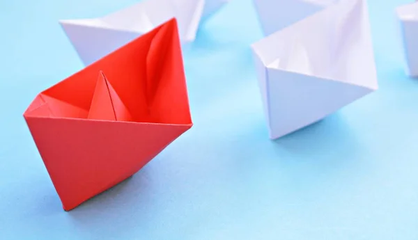 A red self-folded paper boat lies on a blue surface, followed by many white boats - concept symbolizing leadership