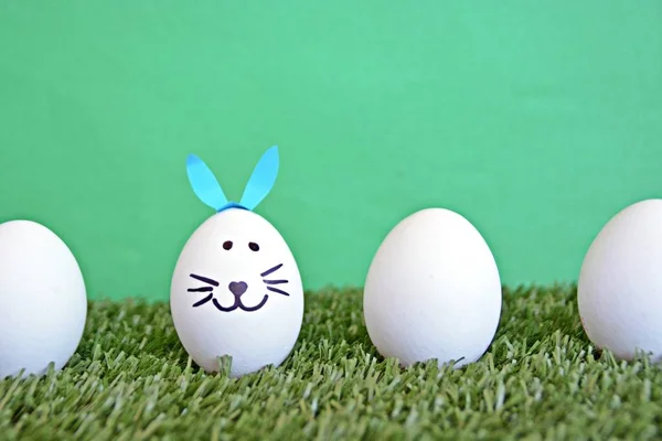 eggs painted with the face of a hare and glued to rabbit ears lie on a grass field in front of a wooden background with space for text or other elements