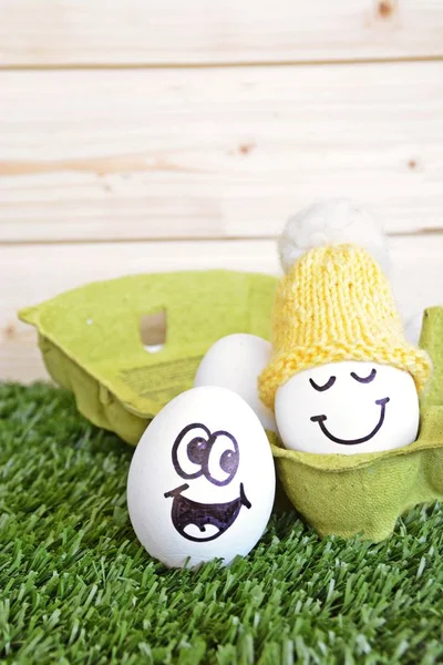 Two eggs with funny faces painted on it lie on a grass field. One of the eggs is in an egg box with other white eggs, the other is next to it