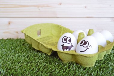 Üzerinde boyalı komik yüzler ile iki yumurta bir çim alanda yalan. Yumurtaların biri diğer beyaz yumurtalar ile bir yumurta kutusunda, diğeri yanında