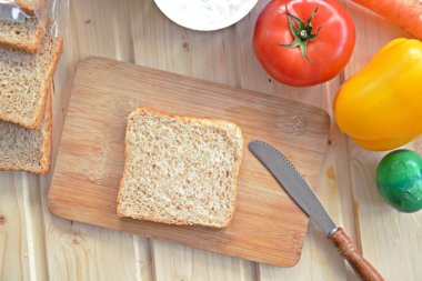 Kaba-taneli krem peyniri ve Cress ile bir kepekli tost bir bıçak yanında bir tahta tahta ve biber ve domates gibi sebze, yanı sıra Cress, salatalık ve havuç bir masada yatıyor