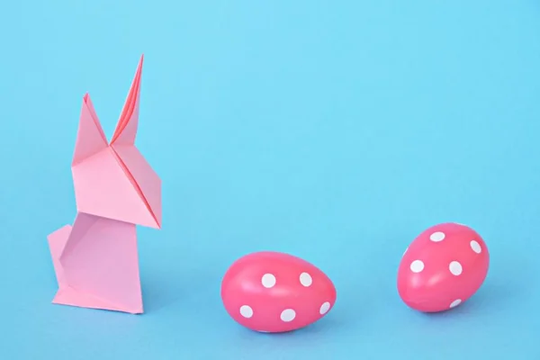 パステルペーパーから折り畳まれたピンクの折り紙のウサギは 白いドットを持つイースターエッグでもある青いベースに立っています 折り紙とテキストやイースターのための他の要素のためのスペースとのコンセプト — ストック写真