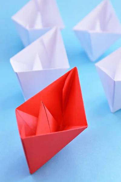 A red self-folded paper boat lies on a blue surface, followed by many white boats - concept symbolizing leadership