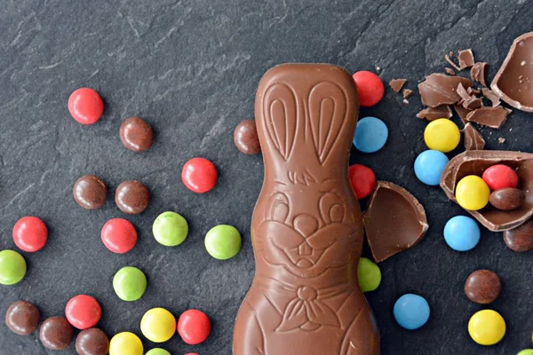 A chocolate easter bunny lies on a dark marble surface with colorful chocolate chips and a few chocolate eggs - concept with sweets for easter with room for text or other elements