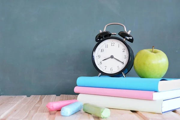 An alarm clock stands in front of a blackboard with colorful books and a green apple - concept with space for text or other elements on the subject of school and time management in elementary school