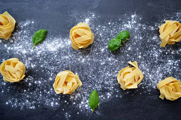 Pasta noodles of the sort tagliatelle lie on a dark marble surface which is lightly sprinkled with flour, next to it are basil leaves - concept as background for fresh prepared noodles