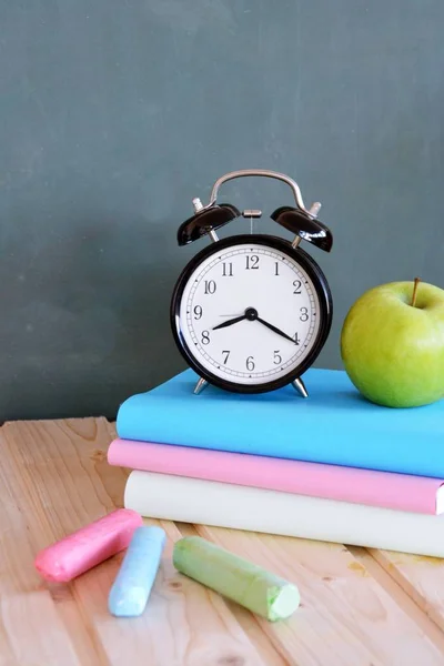 An alarm clock stands in front of a blackboard with colorful books and a green apple - concept with space for text or other elements on the subject of school and time management in elementary school