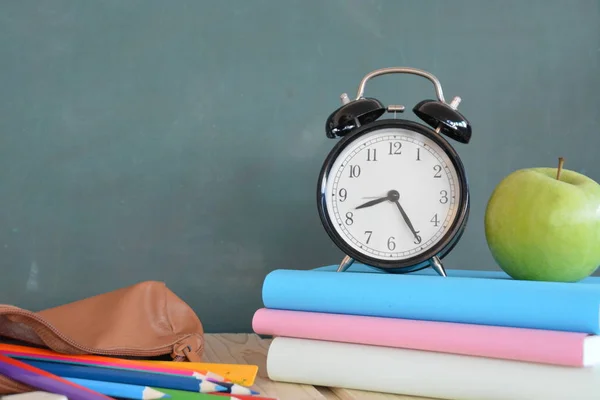 An alarm clock stands in front of a blackboard with colorful books and a green apple - concept with space for text or other elements on the subject of school and time management in elementary school