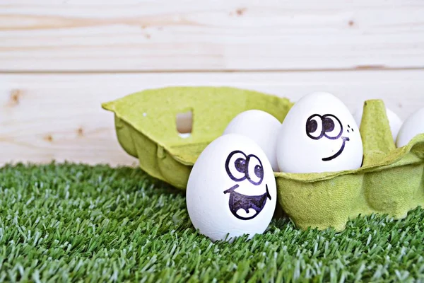 Two eggs with funny faces painted on it lie on a grass field. One of the eggs is in an egg box with other white eggs, the other is next to it