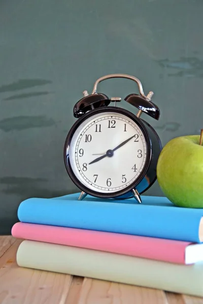 An alarm clock stands in front of a blackboard with colorful books and a green apple - concept with space for text or other elements on the subject of school and time management in elementary school