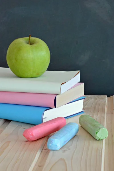 Three books with different colored covers lie on a wooden surface in front of a blackboard with an apple - concept for education and school with place for text or other elements