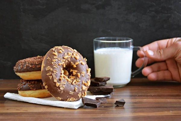 glazed donut with brittle splinters and other chocolate donuts lie on a dark wooden surface against a dark background and a glass of milk - concept for sweet donuts