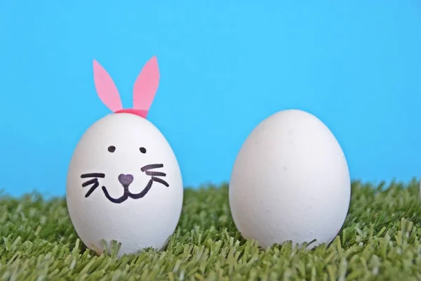 eggs painted with the face of a hare and glued to rabbit ears lie on a grass field in front of a wooden background with space for text or other elements