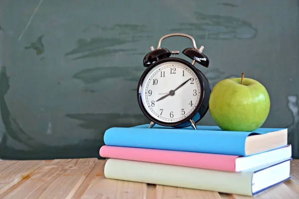 An alarm clock stands in front of a blackboard with colorful books and a green apple - concept with space for text or other elements on the subject of school and time management in elementary school