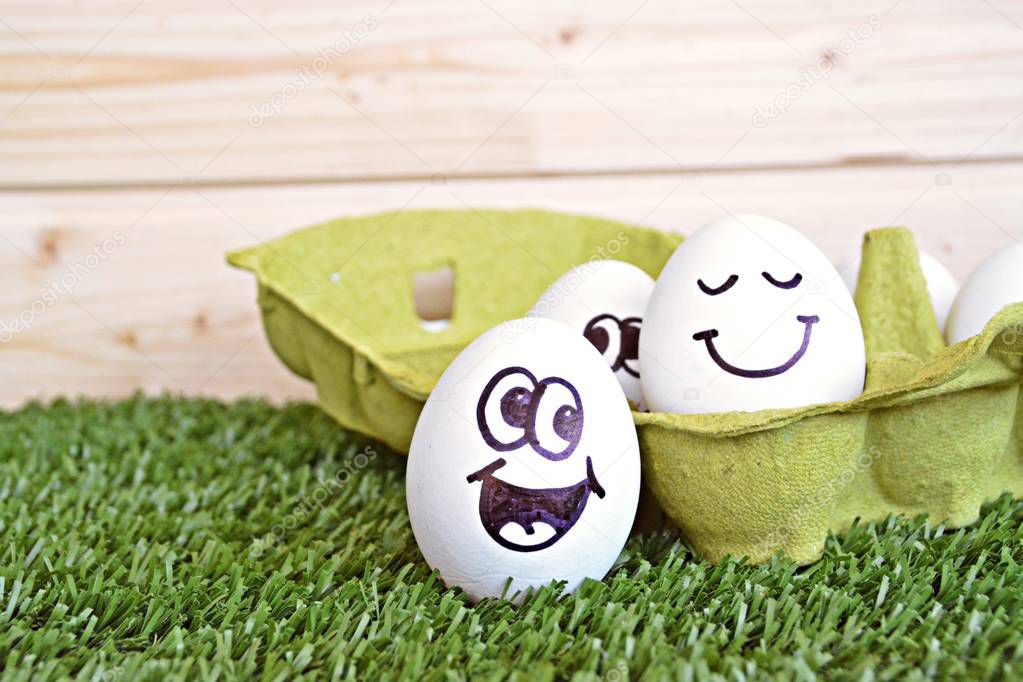 Two eggs with funny faces painted on it lie on a grass field. One of the eggs is in an egg box with other white eggs, the other is next to it