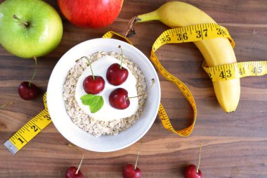 Yulaf ezmesi dolu beyaz bir kase, yanında meyve ile yoğurt ve kiraz bir topak ile, yanında bir bant ölçüsü ile, ahşap bir yüzey üzerinde yatıyor - sağlıklı beslenme için kavram 